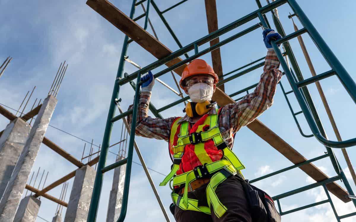 Operario de construcción subiendo por un andamio con equipo de protección individual completo: casco, chaleco de alta visibilidad, protección auditiva y mascarilla